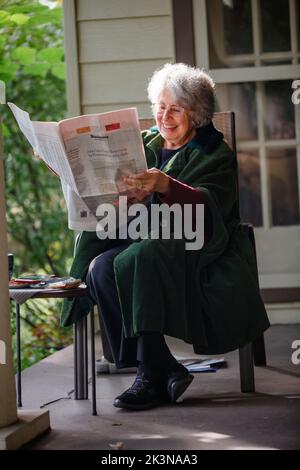 Una donna anziana felice dai capelli grigi siede sul portico leggendo il giornale Foto Stock