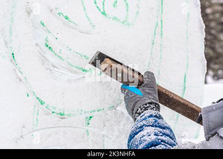 L'artista scultore, utilizzando un coltello in metallo, taglia la figura di ghiaccio di Capodanno dal blocco di ghiaccio secondo lo schizzo finito. Foto Stock