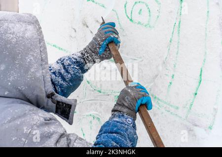 L'artista scultore, utilizzando un coltello in metallo, taglia la figura di ghiaccio di Capodanno dal blocco di ghiaccio secondo lo schizzo finito. Foto Stock