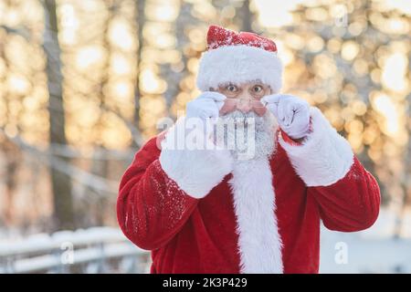 Vita in su ritratto di Babbo Natale tradizionale guardando la macchina fotografica sopra gli occhiali e guardando sorpreso nella foresta invernale all'aperto, copia spazio Foto Stock