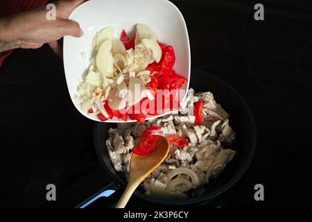 una mano che aggiunge il pomodoro e la cipolla tritati nella carne di pollo friggitrice Foto Stock