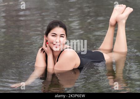 Felice outsize giovane donna in un unico pezzo nero costume da bagno sdraiato e relax in acqua minerale in piscina all'aperto presso spa, terme resort Foto Stock