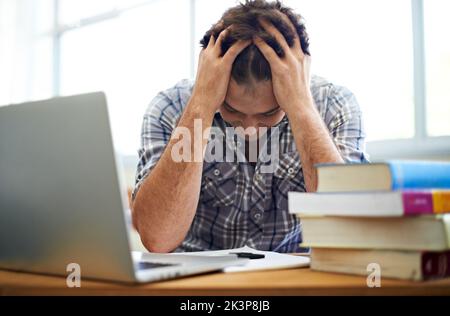 Lo stress dell'esame gli sta arrivando...Uno studente seduto con la testa in mano accanto a un mucchio di libri di testo e al suo computer portatile. Foto Stock