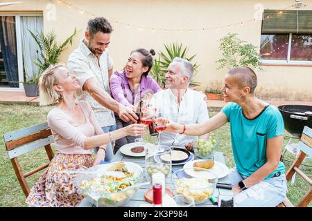 Gruppo di uomini e donne di mezza età che brindano bicchieri di vino durante la cena nel cortile della casa. Amici adulti che si divertono durante il fine settimana. Stile di vita Foto Stock