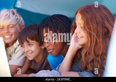 Collegato mentre fuori campeggio. Primo piano di quattro bambini che usano un notebook mentre sdraiati in una tenda insieme. Foto Stock