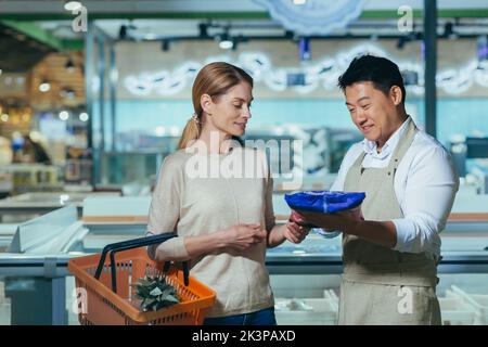 Consulente in un supermercato. Una giovane donna bella vuole comprare prodotti congelati nel negozio. Un consulente dipendente, un uomo asiatico, la aiuta a scegliere. Parlano, sorridono Foto Stock