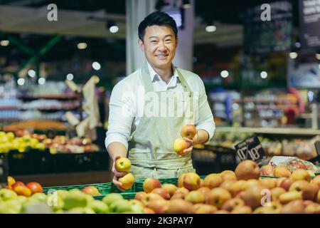 Vita in su ritratto di un bell'uomo asiatico bello che lavora in un supermercato e sorridendo alla macchina fotografica. In piedi in un grembiule di lavoro vicino a verdure e frutta separate. Tiene le mele nelle sue mani. Foto Stock