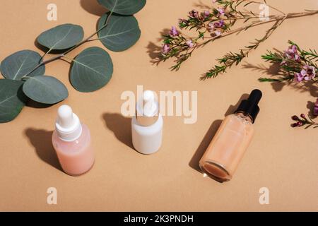 Flaconi contagocce cosmetici, fiori e rami di eucalipto su sfondo beige. Cosmetici naturali, aromaterapia e cura della pelle concetto. Vista dall'alto. Foto Stock