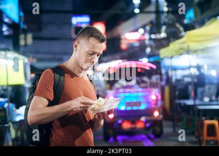 Turista di notte Bangkok. Uomo con zaino mangiare cibo locale al mercato di strada. Foto Stock