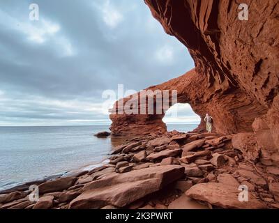 Una donna che cammina sulle rocce rosse a Cavendish Beach sotto il cielo nuvoloso, Prince Edward Island, Canada Foto Stock