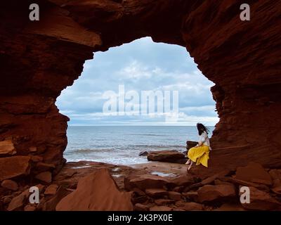 Una giovane donna seduta sulle rocce sotto le scogliere del mare a Cavendish Beach, Prince Edward Island, Canada Foto Stock