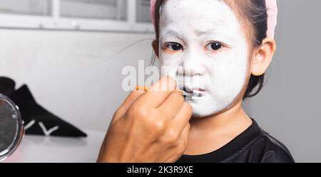 Madre asiatica e figlia capretto ottenere trucco halloween faccia pittura assomiglia a fantasma, ritratto della donna che applica la spazzola di colore alla faccia della ragazza del bambino per pa Foto Stock