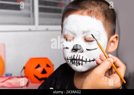 Madre asiatica e figlio di capretto ottenere trucco haloween pittura faccia assomiglia a fantasma, ritratto della donna che applica il pennello di colore al viso del bambino per partito, felice Foto Stock