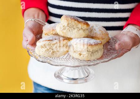 Una donna vestita in modo intelligente è mostrato tenendo freschi scone tradizionali pianura su un piedistallo torta. Le torte sono leggermente spolverate con zucchero a velo Foto Stock