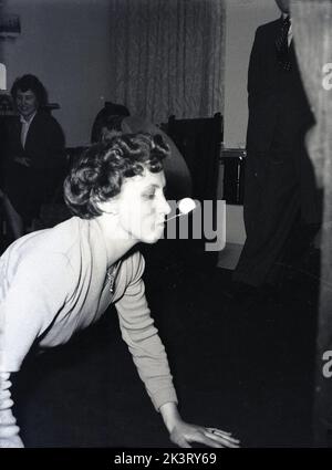 1956, storica Gran Bretagna del dopoguerra, all'interno di una sala d'ingresso, una signora che partecipa a una partita di festa, sul pavimento con le mani e le ginocchia con un cucchiaio in bocca che bilancia una piccola palla, mentre tenta di muoverla, Inghilterra, Regno Unito. Foto Stock