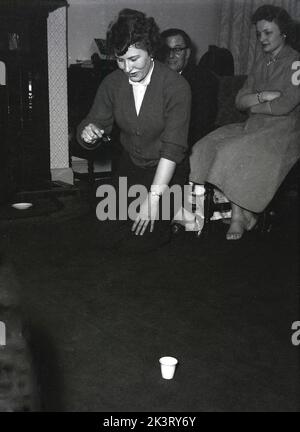 1956, storica Gran Bretagna del dopoguerra, all'interno di una sala d'ingresso e una signora che partecipa a una partita di festa, sul pavimento in ginocchio tenendo una forchetta con una piccola palla rotonda, tentando di farla entrare nella coppa sul pavimento, Inghilterra, Regno Unito. Foto Stock