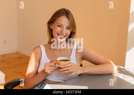 Allegra donna bionda tenere una tazza di caffè vicino notebook sul tavolo a casa, immagine stock Foto Stock