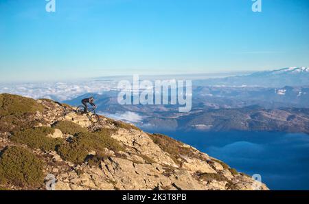 Vista laterale del ragazzo con casco che scende in bicicletta dalla cima della collina e vista pittoresca delle montagne nelle nuvole Foto Stock