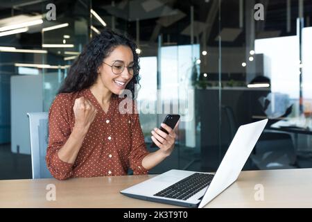 Giovane imprenditrice ispanica di successo che lavora all'interno di un ufficio moderno, una lavoratrice che utilizza un computer portatile, che sorride il telefono e festeggia felice la vittoria e trionfa. Foto Stock