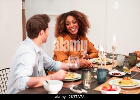 Allegro romantico coppia multirazziale flirting mentre si siede al tavolo e cena con gli amici durante la festa di casa Foto Stock