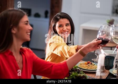 Vista laterale di allegri amici diversi che celebrano e si aggraffano con i vini mentre si siede al tavolo servito con deliziosa cena a casa Foto Stock