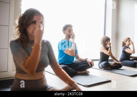 Gruppo di persone concentrate in abbigliamento sportivo seduto con le gambe incrociate e tenendo le dita in movimento Nasarga mudra mentre si pratica esercizio respiratorio Foto Stock
