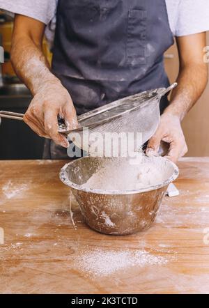 Panettiere maschio anonimo che indossa grembiule in piedi a tavola di legno e setacciare la farina con setaccio metallico in panetteria Foto Stock