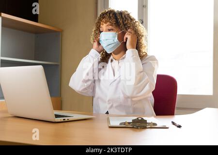 Medica etnica femminile in uniforme mettendo su maschera usa e getta mentre guardando lontano in ufficio Foto Stock