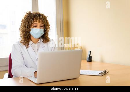 Medica etnica femminile in maschera usa e getta e tipizzazione uniforme su netbook a tavolo in clinica Foto Stock