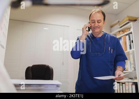 Sorridente medico adulto in uniforme tenendo carta e parlando sul telefono cellulare in sala medica Foto Stock