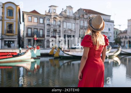 Vista posteriore dell'irriconoscibile turista femminile in sundress e cappello che si erge ammirando il fiume della città in Aveiro Portogallo Foto Stock