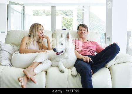 Coppia che riposa sul divano con il cane del pastore svizzero bianco mentre si gode il fine settimana insieme e guardando la TV a casa Foto Stock