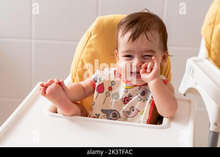 Carino positivo barefooted bambino sorridente e mordente dito mentre si siede in sedia da pranzo in cucina Foto Stock