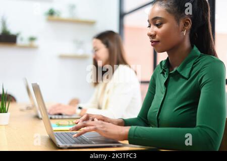 Vista laterale di una dipendente afro-americana concentrata utilizzando un computer portatile, una donna d'affari nera sorridente risponde alle e-mail, una donna freelance che lavora seduta nello spazio di coworking Foto Stock