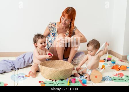 Corpo pieno di felice giovane madre barefooted con i capelli rossi lunghi sorridenti mentre siede sul tappeto molle con i bambini curiosi adorabili che giocano con il giocattolo colorato Foto Stock