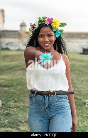 Felicissima donna in corona floreale che raggiunge la mano con fiore daisy blu verso la fotocamera mentre si trova in piedi sul prato verde Foto Stock