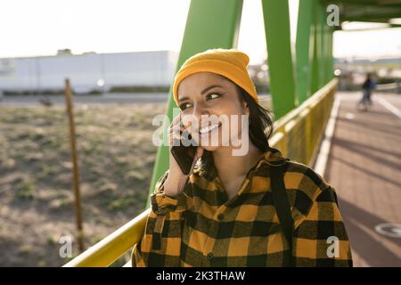 Positiva giovane latino-americana con lunghi capelli scuri in elegante camicia a scacchi in piedi sul ponte e telefono cellulare parlante nelle giornate di sole Foto Stock