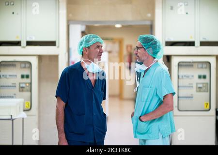 Vista laterale di due uomini in scrub che parlano l'uno con l'altro mentre in piedi in teatro operatorio dopo l'intervento chirurgico Foto Stock