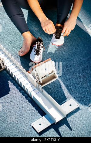 Dall'alto l'anonimo sportswoman corto indossa le scarpe da pista mentre si siede sulla pista vicino ai blocchi di partenza prima di correre sullo stadio Foto Stock