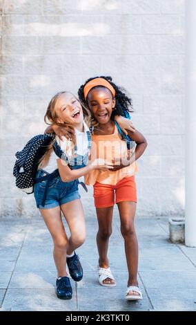 Allegra ragazza afroamericana con la migliore amica che si diverte e cammina in abbraccio con zaini Foto Stock