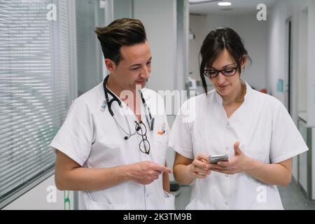 Coppia di pediatri in uniforme bianca con telefono che cammina lungo il corridoio della clinica dopo la fine del turno di lavoro Foto Stock