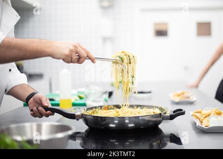Crop uomo in giacca bianca chef cattura spaghetti dalla padella mentre si è in piedi in cucina ristorante Foto Stock