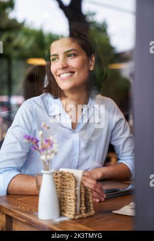 Ritratto girato attraverso la finestra di una donna sorridente latino-americana seduta al caffè Foto Stock