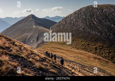 Vista posteriore ad angolo alto di un gruppo di viaggiatori con zaini che camminano su un sentiero stretto tra montagne rocciose con pendici erbose in estate a Pyr Foto Stock