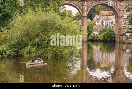 Una barca a remi su un fiume con un viadotto storico dietro. Gli alberi sono sulla riva e gli archi si riflettono nell'acqua. Le case sono sullo sfondo. Foto Stock