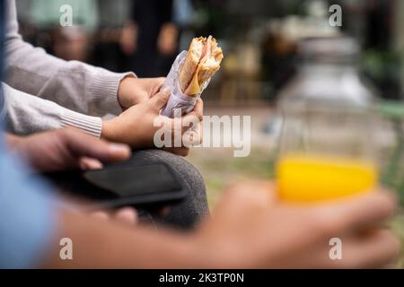 Primo piano di una mano maschile fuori fuoco che tiene una bottiglia di succo d'arancia e un paio di mani che tengono un panino Foto Stock