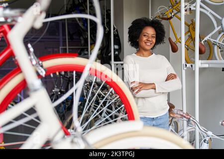 Scatto medio di una donna afroamericana proprietaria di un negozio di biciclette nel suo negozio con biciclette in primo piano Foto Stock