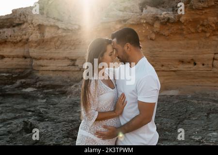 Vista laterale di un uomo allegro che abbraccia donna incinta in vestito mentre si trova contro la scogliera di mattina in natura Foto Stock