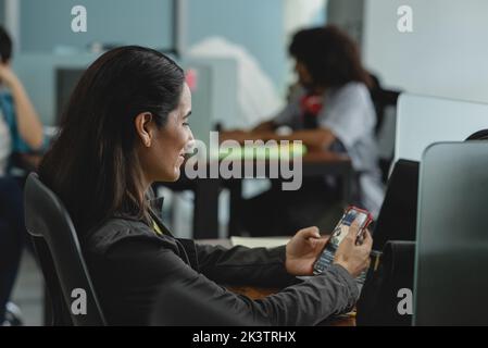 Vista laterale di una donna ispanica sorridente con capelli scuri in abiti casual seduti a un tavolo e navigando in rete sociale su smartphone durante la pausa in offic Foto Stock