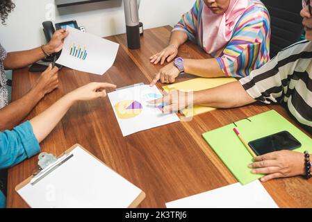 Da sopra gruppo di colleghi multirazziali penosi in abiti casual seduti a un tavolo di legno con carte e discutere i dettagli delle tabelle nello spazio di lavoro Foto Stock